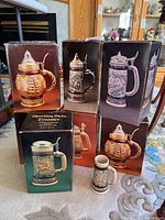Photo showing six decorated ceramic steins in original boxes featuring different designs including sailing ships, western themes, and sporting crests.