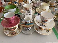 Photo showing a group of teacups and saucers stacked and arranged, visible different colors and patterns including pink, green, white with fruit and floral motifs, including a miniature souvenir cup.