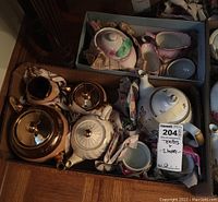 Two boxes containing various teapots, creamers, and sugar bowls with floral and gold designs packed with wrapping papers.