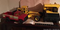 Three Tonka toys on a wooden surface: large dump truck, scraper, and red pickup truck with ruler for scale
