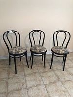 Front view of three black wooden bistro chairs with curved backrests and fabric-covered seats arranged on a tiled floor against a plain wall.