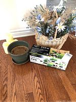 Wicker basket filled with blue and white silk flowers and dried foliage, green ceramic flower pot, boxed set of personalized plant pots, and two yellow battery operated candles displayed on wooden floor.