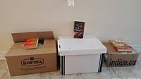 Three boxes on floor, two cardboard boxes and one white storage box, with some books placed on top and on floor in front, showing range of novels