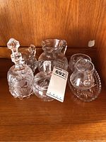 Five clear crystal pieces including a bell with stopper, pitchers, and bowl on a wooden shelf