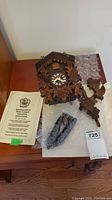Complete cuckoo clock with carved leaf motifs, Roman numeral clock face, original owners manual, pinecone-shaped weights, and extra carved wooden pieces, on wooden surface