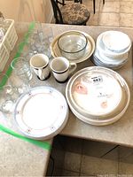 Overview of stacked dishes, bowls, plates, mugs, and glasses on countertop showing Noritake and Royal Doulton pieces with some clear glassware.