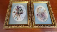 Two framed pressed flower artworks side by side showing detailed floral arrangements and matching ornate gold frames.