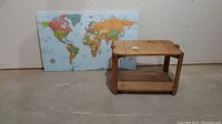 World map mounted on board alongside a two-tier wooden table in a plain setting.