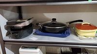 Shelf with cooking items including a speckled enamel roasting pan, black non-stick frying pan with glass lid, a yellow enameled pot with orange lid, and a glass casserole dish.