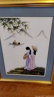 Full view of framed silk painting depicting two women near boat and bamboo, in gold frame with blue mat.