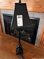 Black metal table lamp with square black fabric lampshade seen from front against a fireplace background.
