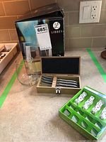 Nine stemless wine glasses in front of Libbey box, wooden box with five cocktail spreaders, and a set of dice in green box.