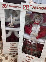 Pair of two vintage 28 inch tall animated and illuminated Christmas figures in their original white cardboard boxes with '28" Motion-ed Animated and Illuminated Christmas Figure' labeling and plastic windows.