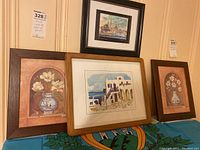 Four framed pictures: three floral images with white flowers in vases, one small landscape of Greek-style buildings by the sea, and one detailed cityscape of mosques by a waterfront, all framed and shown together on a patterned cloth.