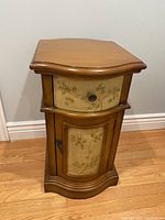 Front view of small wooden cabinet showing painted floral design on drawer and door, metal pulls, and curved wood edges.