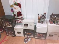 Wide shot showing multiple boxed Christmas village houses including Dickens Keepsake and other ceramic homes, with Santa Claus mailbag decoration behind them.