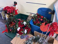 Photo showing multiple Christmas wreaths with red and white flowers, pine cones, and artificial greenery along with poinsettias and decorative items on the floor and in tubs.