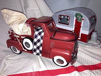 Photo showing vintage style red metal pickup truck with plaid side panel detailing and beige sack in the truck bed, with camping trailer visible in background.