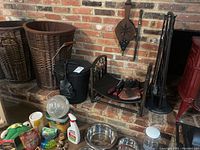 Overview photo showing some fireplace accessories on brick hearth including wicker baskets, ash bucket, gloves, wood holder, and some household items in front.