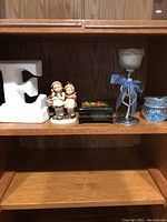 Full view of items on shelf including white ceramic letter 'E', Hummel figurine, black keepsake box, rose candle holder with ribbon, and blue glass jar.
