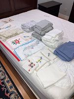 White, grey, blue bed linens and embroidered table linens and napkins arranged on bed