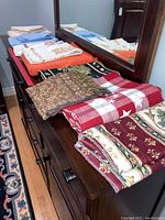 Overview of assorted linens including tablecloths and bed scarves on wooden dresser with mirror.