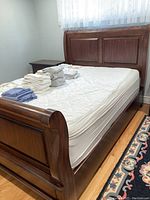 Full view of sleigh bed frame with mattress showing curved headboard and footboard with paneled design. White sheets and folded linens on mattress.