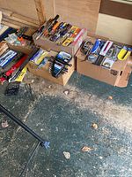 Overall view of multiple cardboard boxes containing hand tools including saws, screwdrivers, clamps, and wrenches, arranged on a floor.