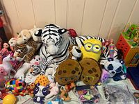 Overview of large stuffed animals including a white tiger, panda, Minion plush, cookie-shaped plush and other assorted plush toys, small figures, and My Little Pony dolls laid out on the floor against a wall