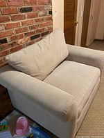 Beige upholstered wide armchair with broad arms and a detachable back cushion, placed next to a brick fireplace.