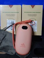 Two UNIGEAR rechargeable hand warmers in pink along with original packaging, wrist strap, and carrying sleeve on a blue background.