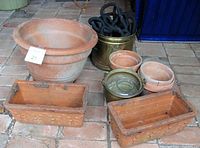 Wide view showing assorted terra cotta and brass pots, including large round cracked terra cotta pot, two rectangular Italian terra cotta pots, two smaller round pots, and brass pots with hose inside.