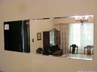 Wall mirror hung on a light-colored wall reflecting a dining room with furniture and drapes, showing its reflective clarity and condition.