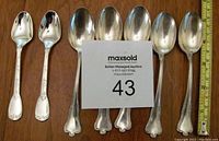 Seven silver teaspoons laid out on a wooden surface, including five of one design with three-lobed handles and two of another design with square bowls and floral handles. A measuring tape shows spoons about 5-6 inches long.