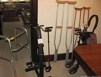 Photo showing two aluminum walkers, Nexus wheeled walker with seat, two pairs of crutches, two canes, and black exercise bike in corner