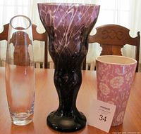 Front view of three vases displayed on table: tall purple amethyst vase, pink floral ceramic vase, and clear glass vase with handle cutout and angular top