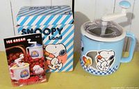 Snoopy and Woodstock themed vintage ice cream maker in light blue with white lid and hand crank, original Snoopy Donvier Land box with blue and white stripes, and printed instructions.