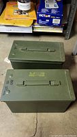 Two green metal ammo boxes placed on concrete floor, showing top side with handles and one with printed caliber details.