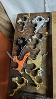 Overview photo of 9 archery thumb releases on a brown surface, showing variation in materials and colors, including brass, silver, black metal, and orange plastic.