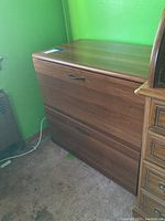 Full view of the two-drawer wooden filing cabinet with visible black handles and brown finish, positioned in a green room corner.