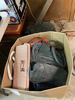 Box filled with multiple women's handbags and purses showing a beige quilted clutch and black leather handbag on top