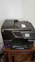 Front and top view of black HP Officejet Pro 8600 printer sitting on wooden furniture showing model label and paper tray.