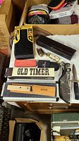 Overall lot with knives in boxes, sheaths, and cases displayed on a cloth surface.