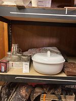 Shelf holding white serving bowl with lid, glass jars with metal lids, cloth placemats with floral pattern, and brown bakeware.