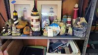 Wide view of metal shelving unit holding multiple lighthouse figures, two framed lighthouse pictures behind, wooden clogs, and jar with lighthouse painting. Items arranged close together, showing size variation and diversity of lighthouse-themed decor.