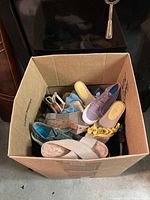 Top-down view of box full of mixed women's shoes showing various styles and colors including purple slip-on sneakers and yellow floral flats.