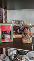 Photo showing three items including Budweiser anniversary stein in box, Norman Rockwell ceramic decanter with people at table scene, and Bavarian-themed ceramic music box stein.