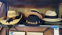 Five hats on a metal shelf: one light tan woven straw hat with black bow band, one black felt cowboy hat with antler decoration, one tan felt brim hat with dark band, one woven straw hat with feather decorations, and one light cream felt brim hat.