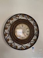 Front view of the large round metal wall clock mounted on the wall showing decorative leaf and vine metalwork around the clock face with Roman numerals.