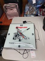 Front view of Snap-On themed square wall clock with red and black hands on white face showing Snap-On logo and tool graphics.